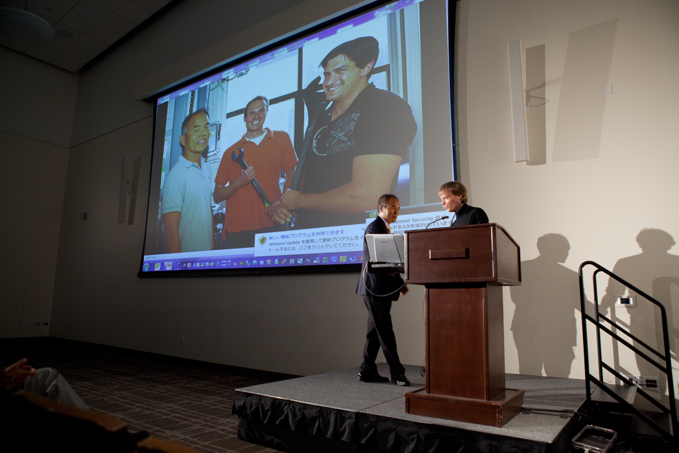 Aumann Lecture 2 - (Two people standing at a podium in front of a projector with image of three people)
