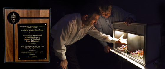 Two people standing in front of two boxes analyzing the two different lights with an award to the left