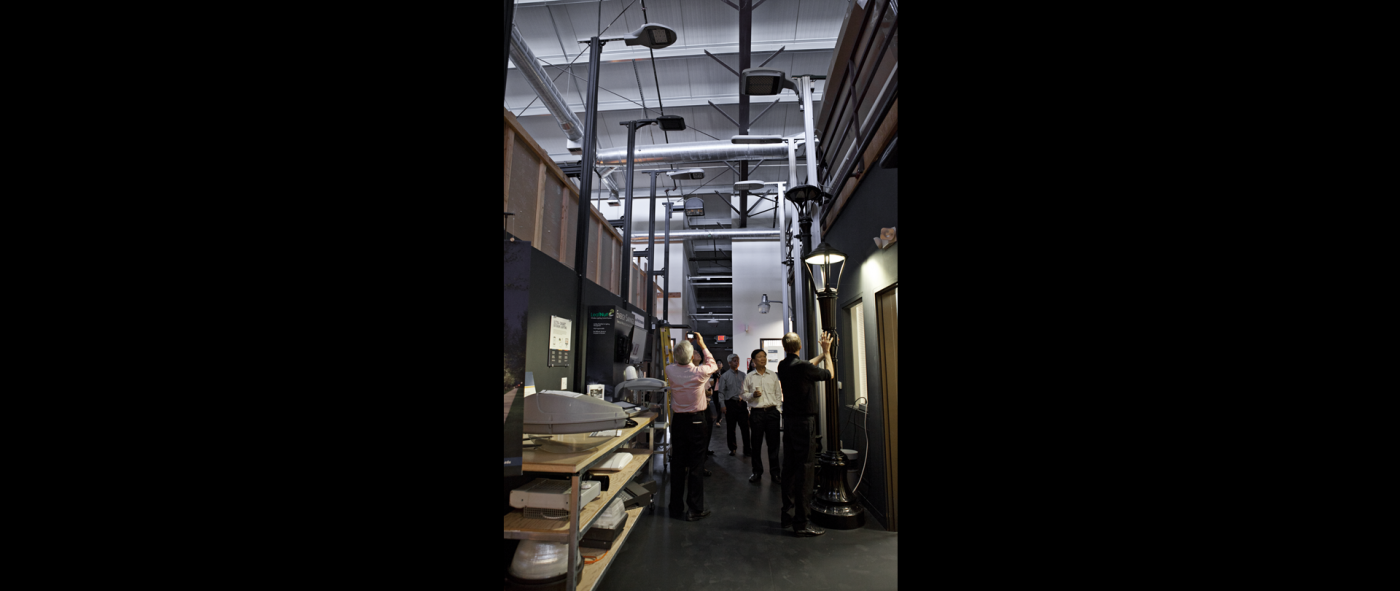 A group of five people looking at different types of exterior light posts lined up inside of a building