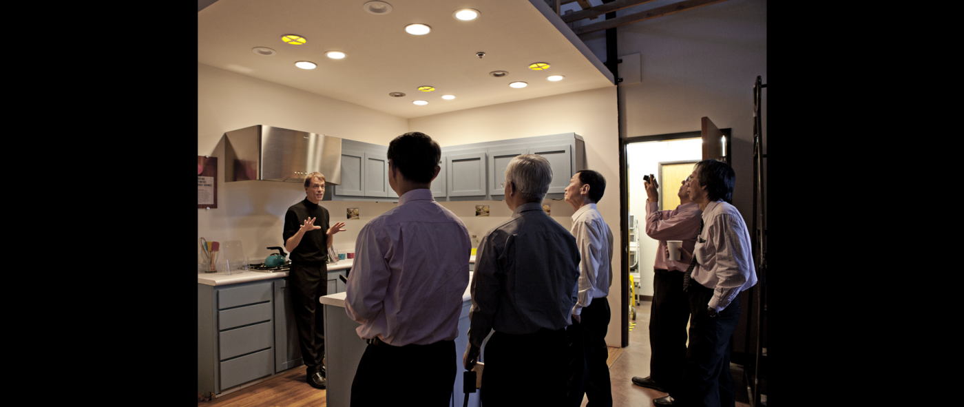  A person standing and talking in front of five other people in a kitchen