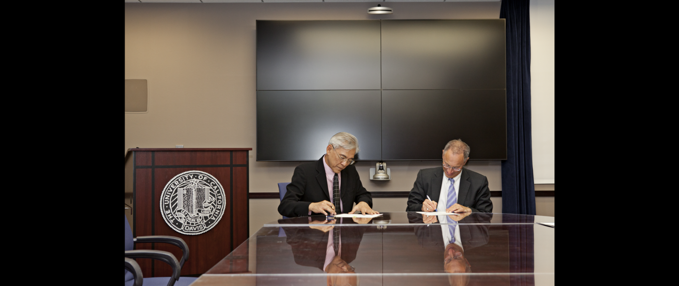 Two people wearing suits and sitting at a conference table, signing formal documents