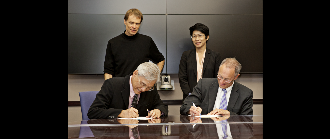 Two people wearing suits and sitting at a conference table signing formal documents with two people standing behind them
