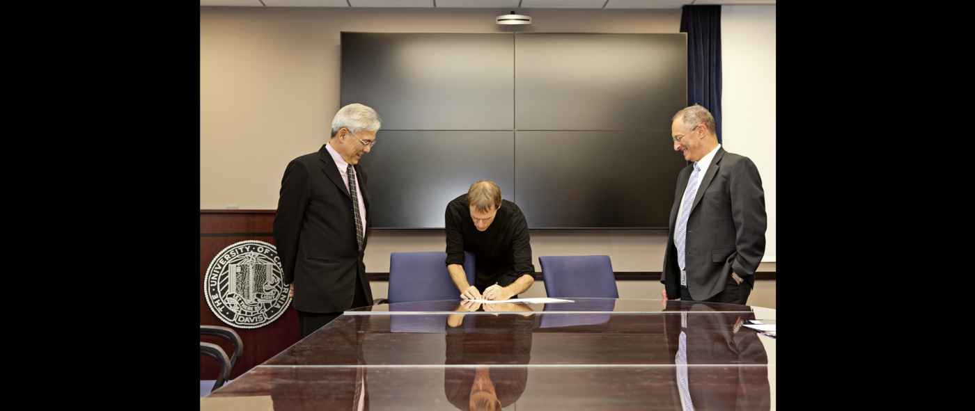 A person in formal attire sitting at a conference table signing formal documents with two people standing on either side