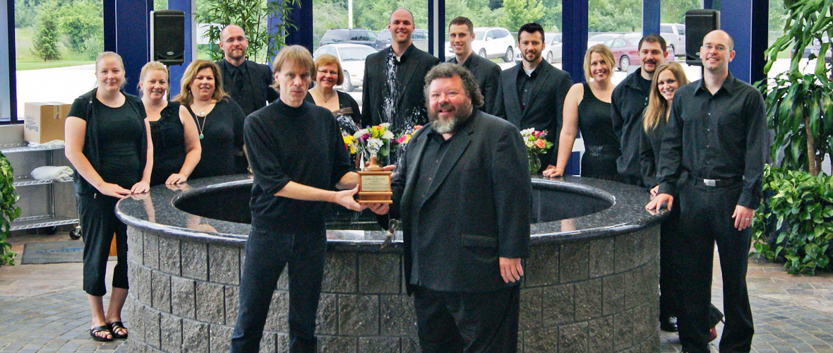 A group of 14 people standing around a circular fountain with two people in the front holding an award