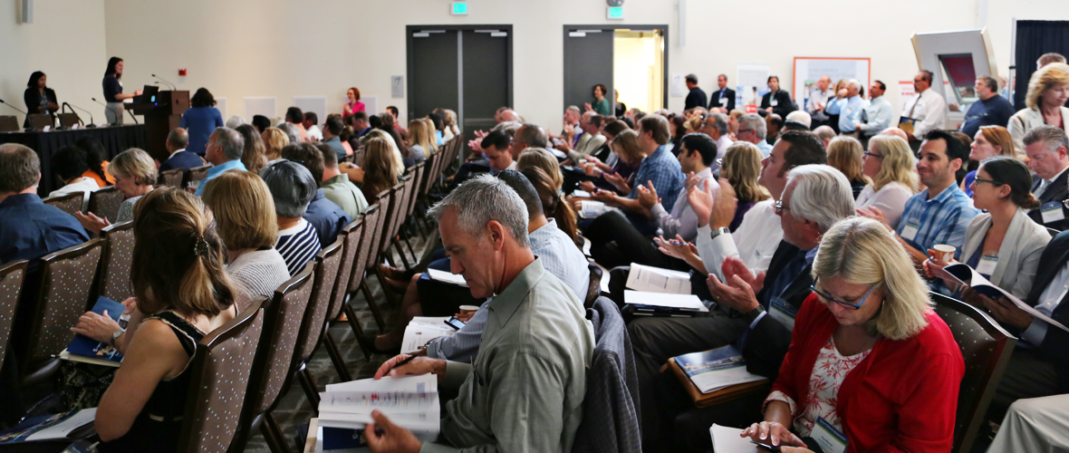 A room full of people sitting in theater seats with name tags and books on their laps. There is a speaker at the front of the room standing at a podium.