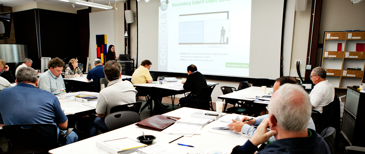 A classroom of multiple people sitting at desks with one person at the front of the classroom talking in front of a slide presentation on a projector screen.