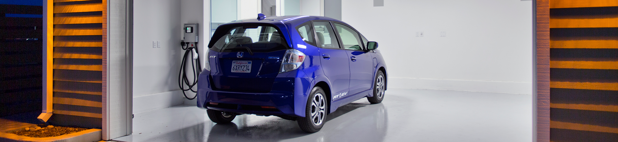A blue Honda Fit being charged electrically inside a two-car garage with white walls