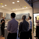  A person standing and talking in front of five other people in a kitchen