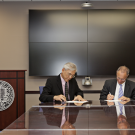Two people wearing suits and sitting at a conference table, signing formal documents