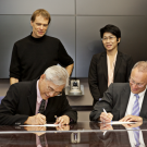 Two people wearing suits and sitting at a conference table signing formal documents with two people standing behind them