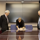A person in formal attire sitting at a conference table signing formal documents with two people standing on either side